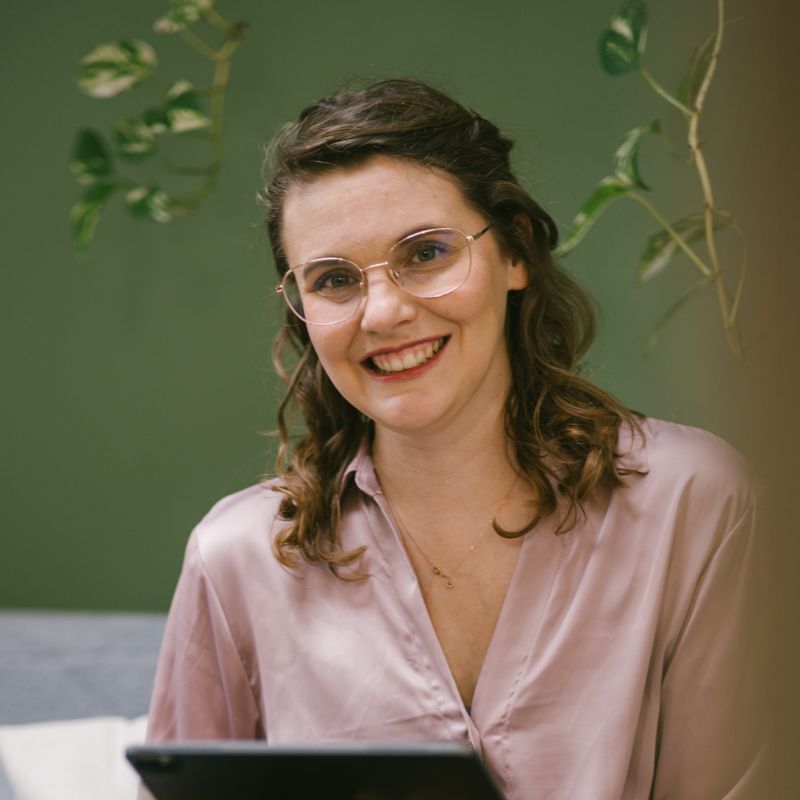 Frau mit lockigem Haar und Brille, die in einem Innenraum mit einem Tablet lächelt. | Mave Consulting