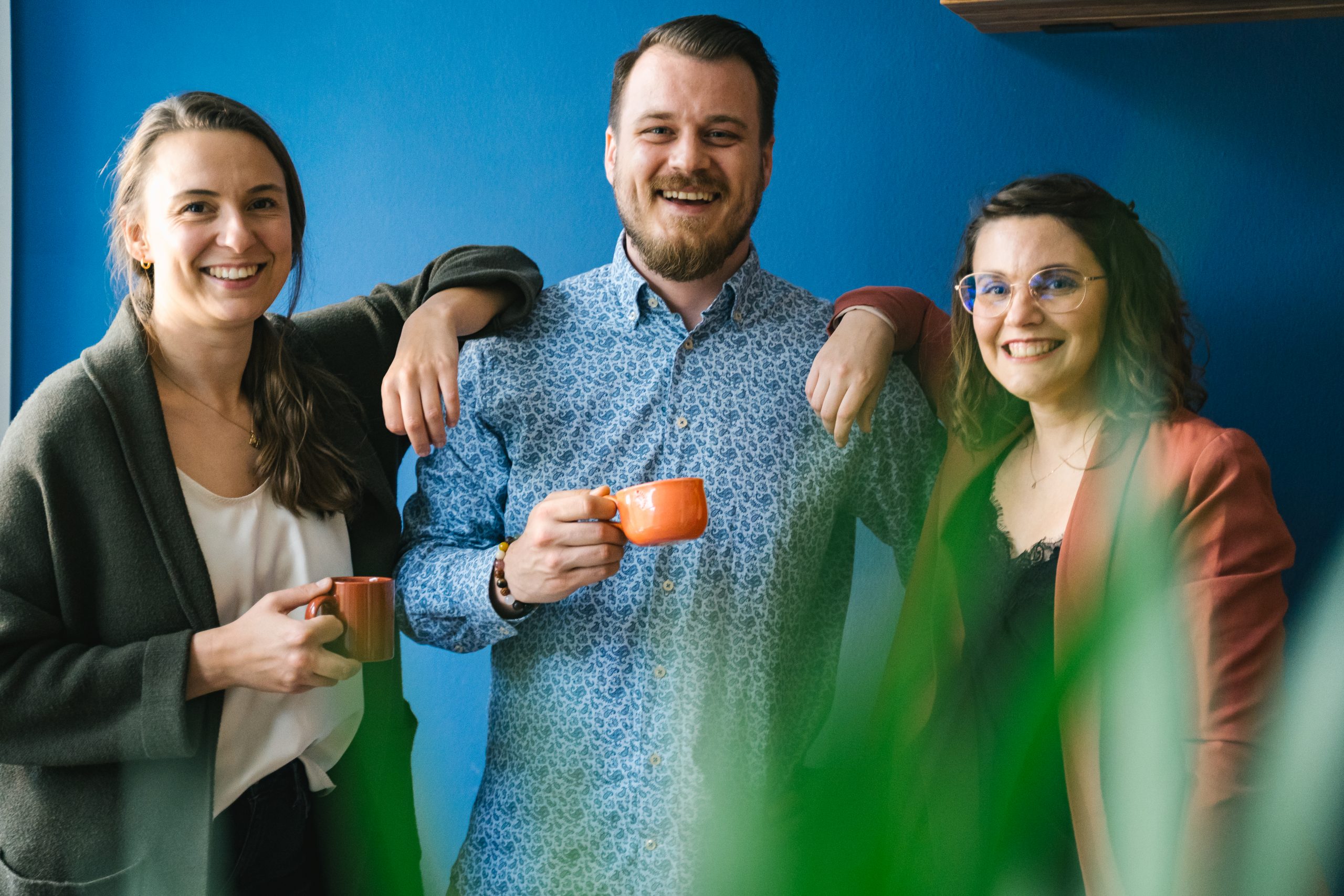 Drei Personen stehen zusammen und halten Tassen in der Hand vor einer blauen Wand. | Mave Consulting