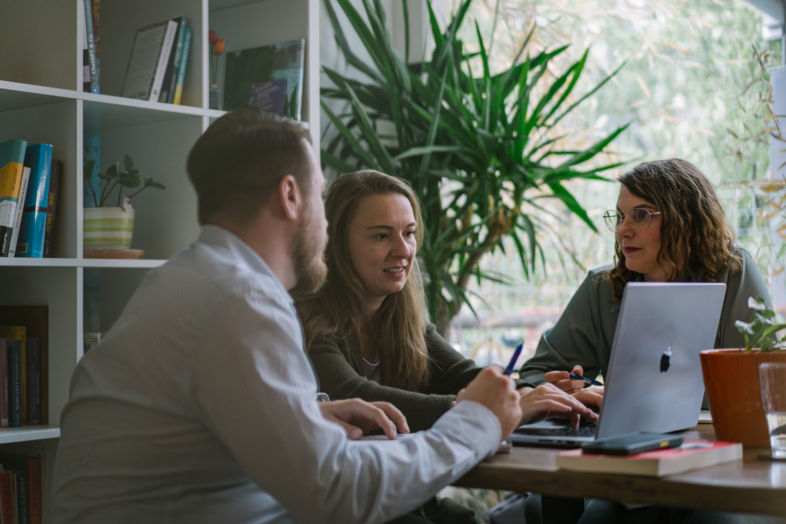 Drei Personen sitzen an einem Tisch, während sie an einem Laptop arbeiten und miteinander sprechen. | Mave Consulting