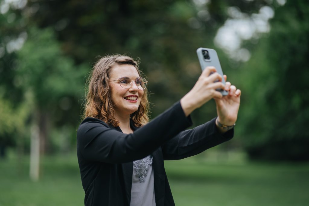 Frau mit lockigem Haar und Brille, die ein Smartphone im Freien hält und lächelt. | Mave Consulting