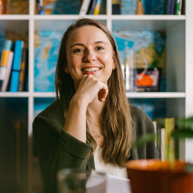 Eine Frau mit langen Haaren lächelt und stützt ihr Kinn auf die Hand in einem Raum mit Regalen. | Mave Consulting